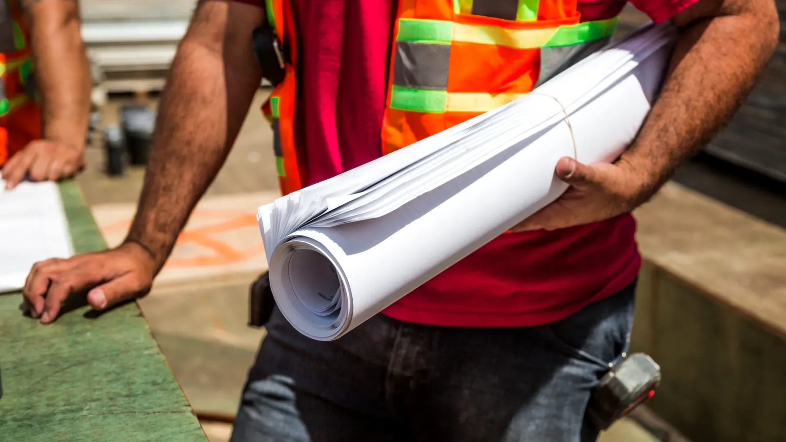 Trabajador con chaleco reflectivo sostiene planos enrollados en una obra en construcción
