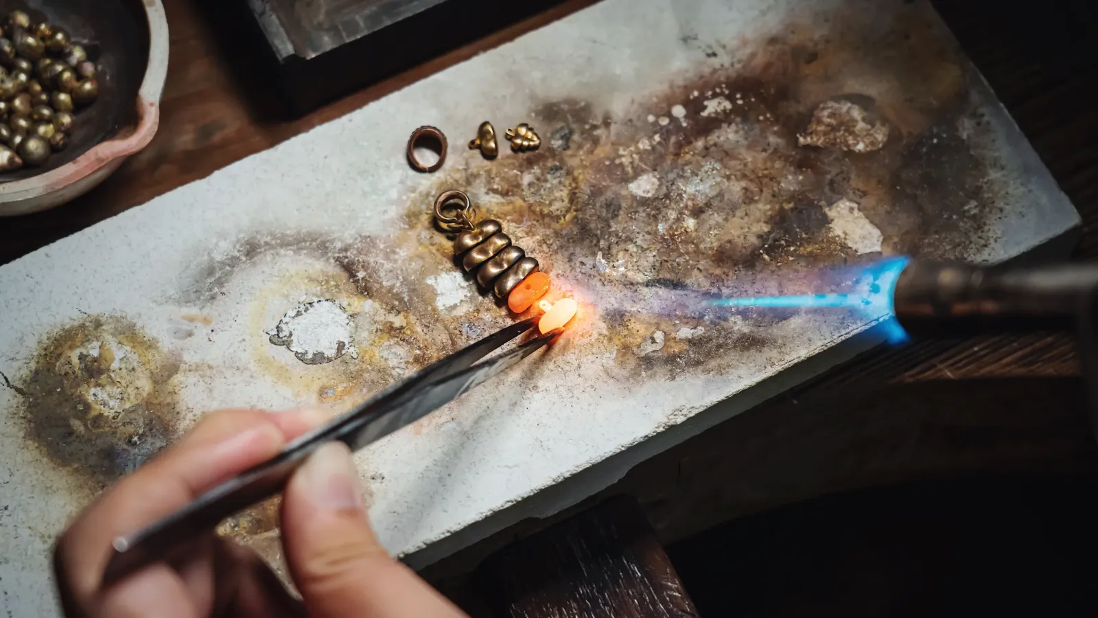 Goldsmith's hands solder a gold-colored piece with a torch on the workbench