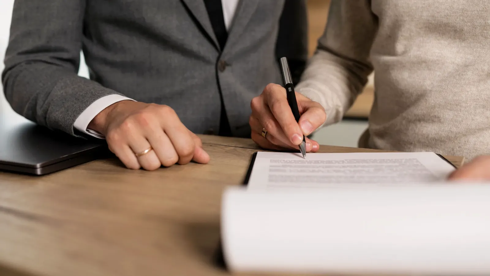 Persona firma un documento sobre una mesa de madera junto a un funcionario de traje gris