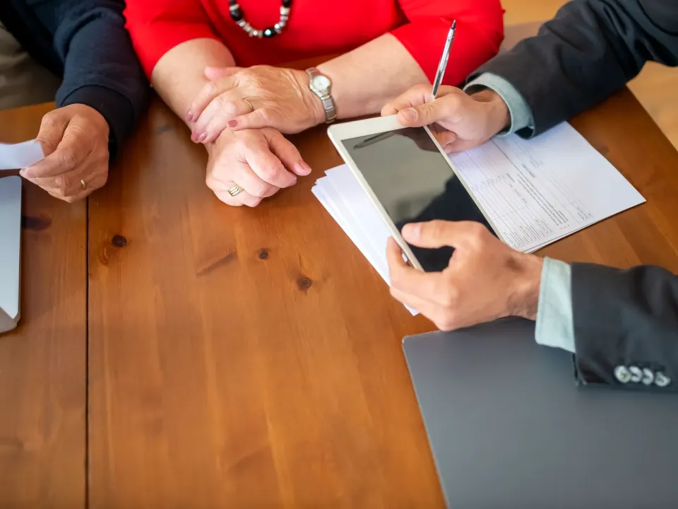 Asesor con tableta y una solicitud impresa atiende a dos socios sentados al otro lado de la mesa