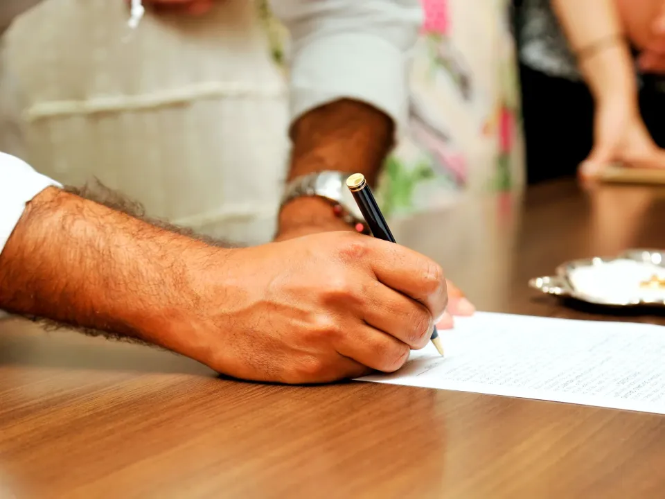 Mano firmando un documento sobre una mesa de madera, con otras personas de pie al fondo