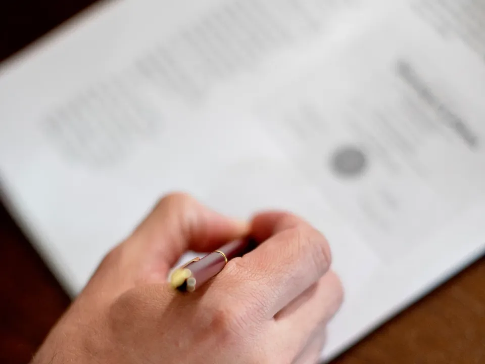 Mano con pluma a punto de firmar un documento formal que lleva un sello impreso