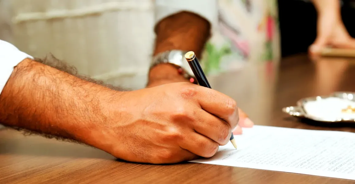 Hand signing a document on a wooden table, with other people standing in the background