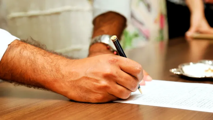 Mano firmando un documento sobre una mesa de madera, con otras personas de pie al fondo